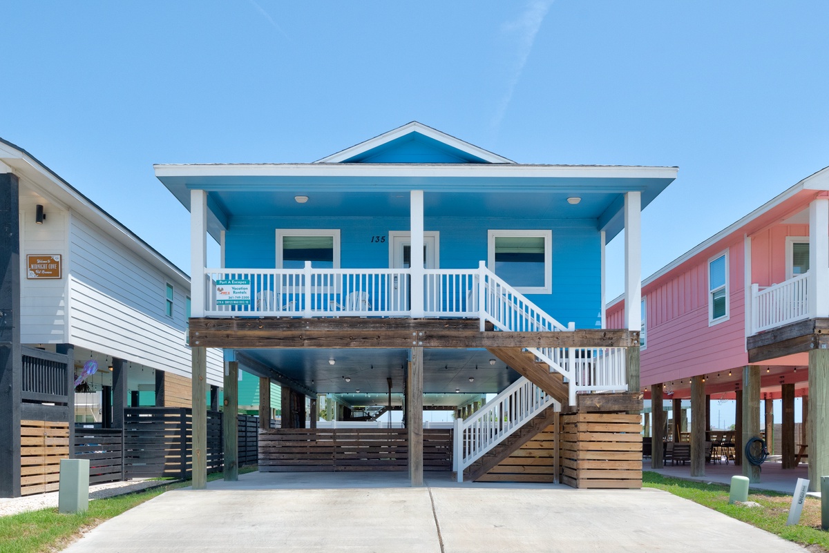 A coastal home elevated on sturdy stilts featuring a bright turquoise facade and welcoming covered porch. The property offers convenient ground-level parking beneath the main living spaces.
