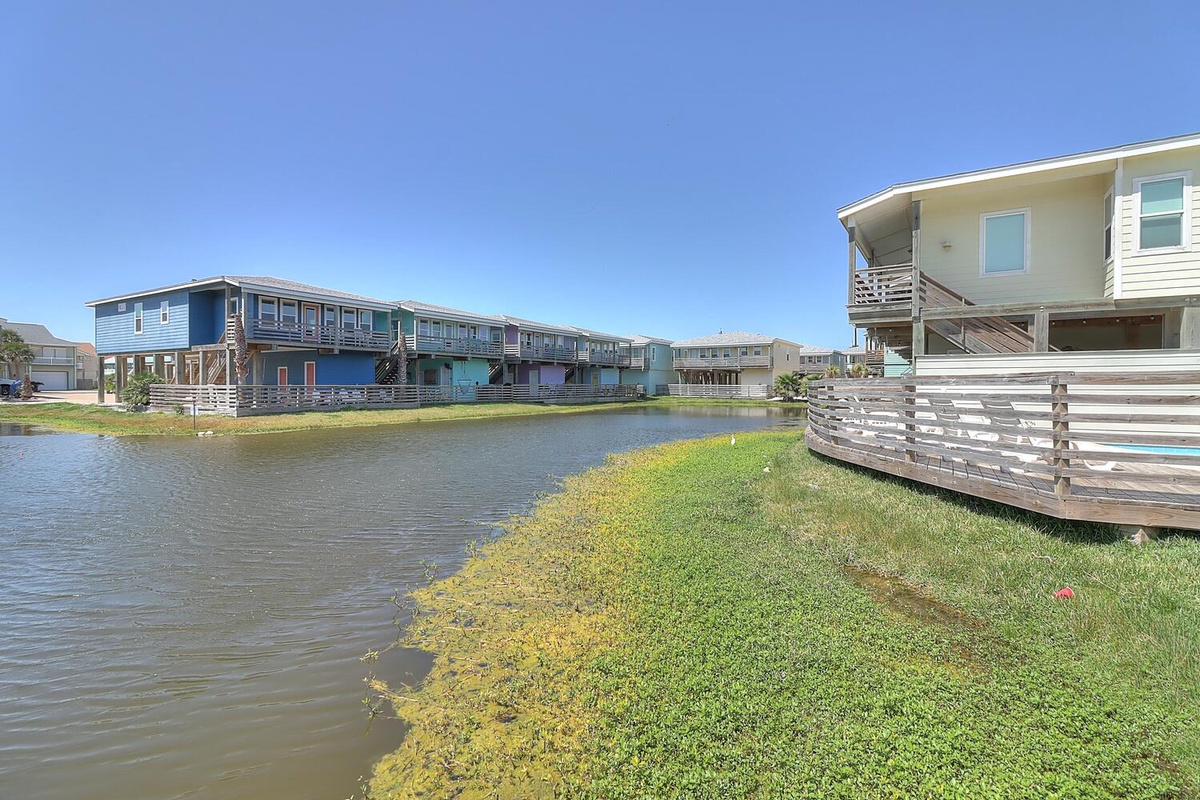 Colorful beach houses line a peaceful waterway with expansive green lawns and elevated decks throughout the coastal community.
