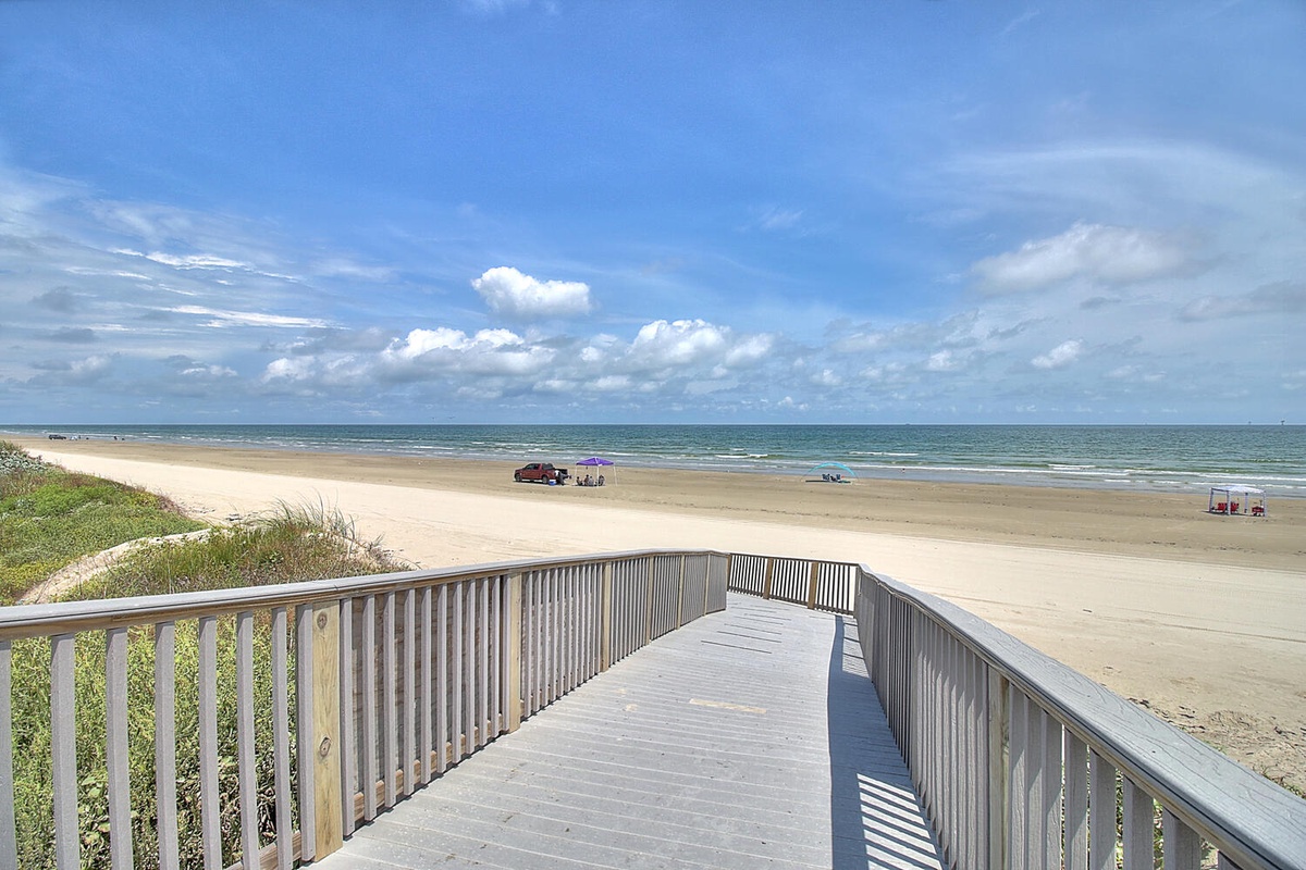 Wide sandy beach stretches endlessly under bright blue skies, with convenient boardwalk access leading directly to pristine coastal waters.