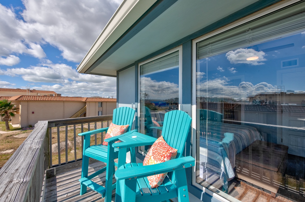 Unwind on your private balcony with vibrant turquoise chairs and decorative pillows, overlooking peaceful neighborhood rooftops under expansive blue skies.