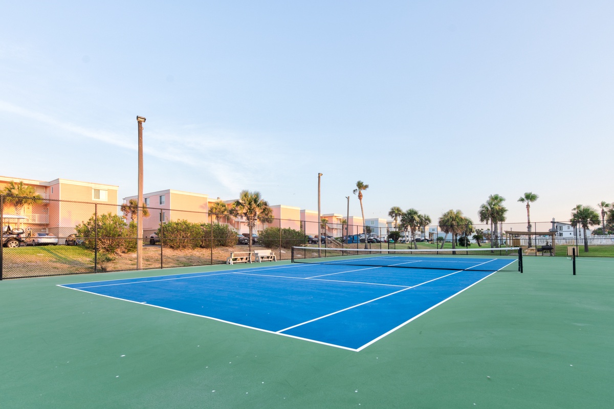 Professional tennis court with blue playing surface surrounded by palm trees and colorful resort buildings.