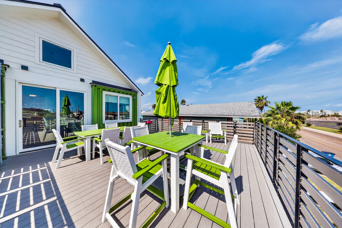 A sunny coastal balcony with cheerful green and white outdoor furniture overlooks palm trees and the surrounding neighborhood.
