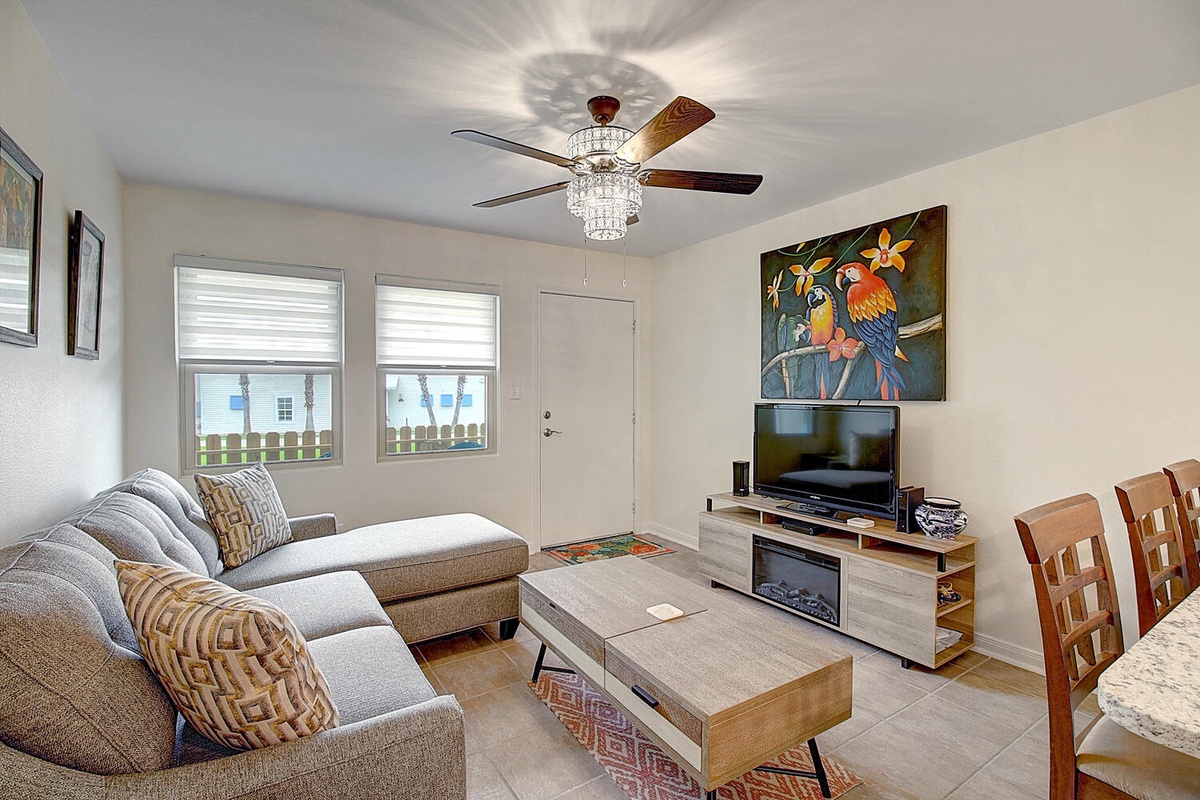 Relax in your bright, modern living space with plush seating, vibrant tropical art, and cozy electric fireplace for perfect comfort.