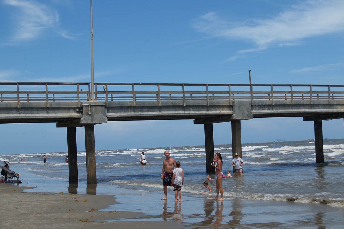 Sandy beach beneath a fishing pier where families enjoy ocean waves and coastal activities under sunny skies.