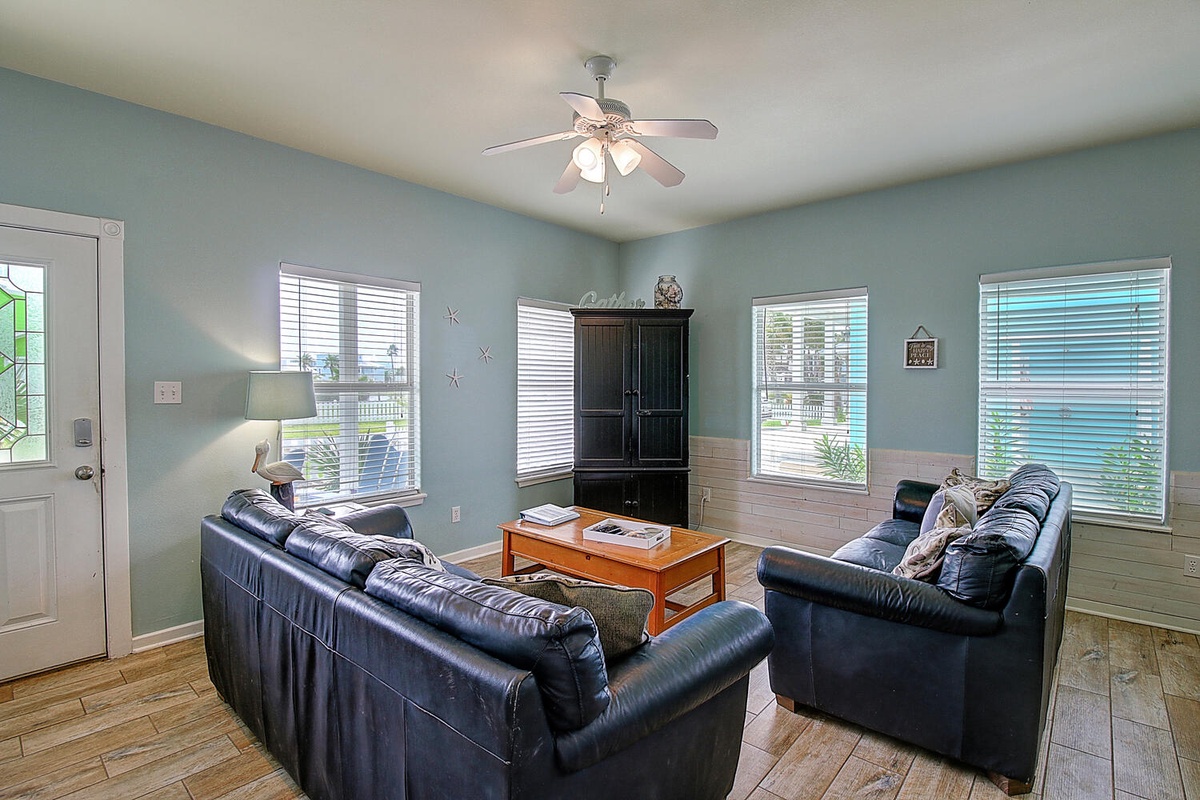Relax in your spacious living room with comfortable leather sofas, ceiling fan, and plenty of natural light streaming through multiple windows.
