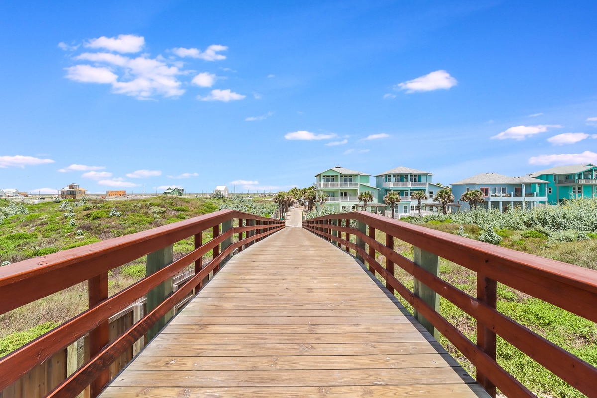 Wooden boardwalk leads through coastal dunes toward colorful beachfront homes under bright blue skies.