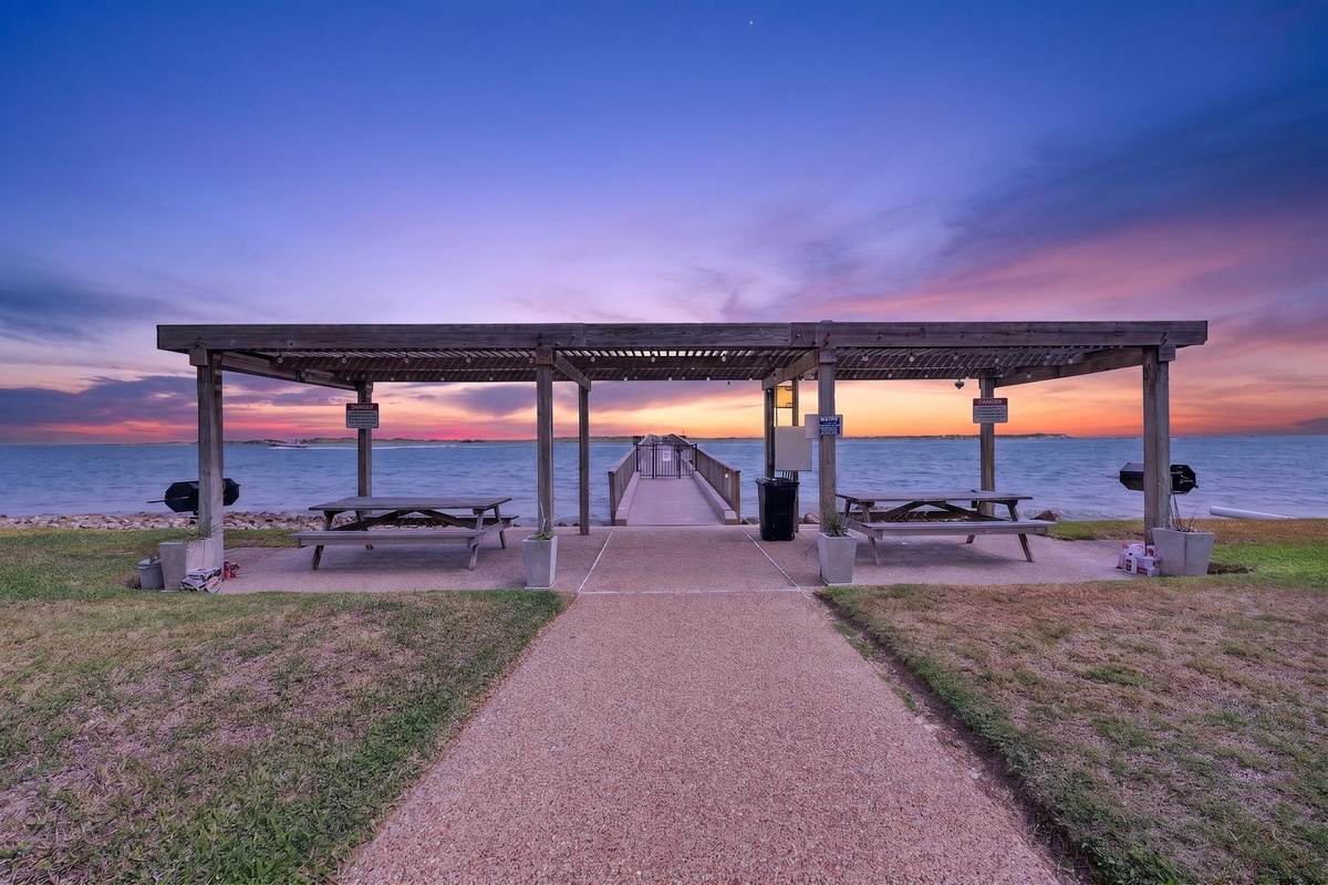 A waterfront pavilion with picnic tables faces a stunning sunset over calm waters, offering a peaceful spot to relax and take in the colorful evening sky.