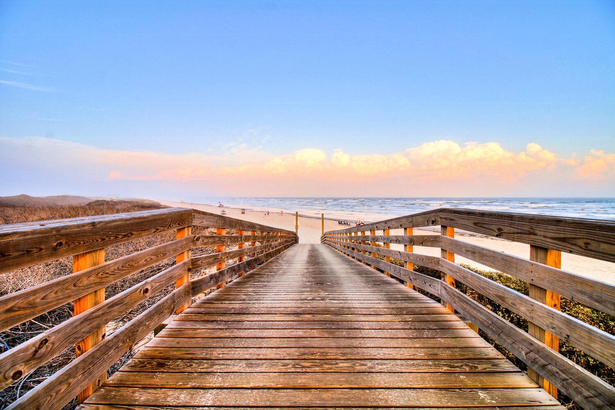 A wooden boardwalk leads toward pristine coastline under golden evening light, with ocean waves and sandy dunes stretching into the distance.