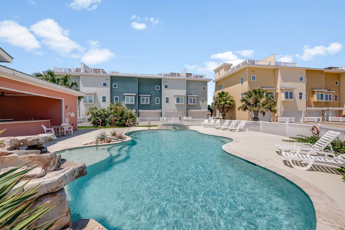 Sparkling community pool surrounded by colorful coastal homes with palm trees and blue skies overhead.