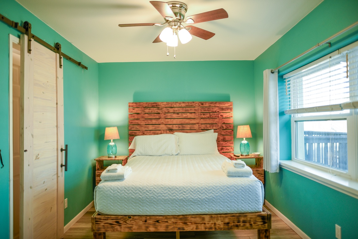 Your peaceful bedroom sanctuary awaits with vibrant turquoise walls, rustic wood furnishings, and soft natural light streaming through the window.