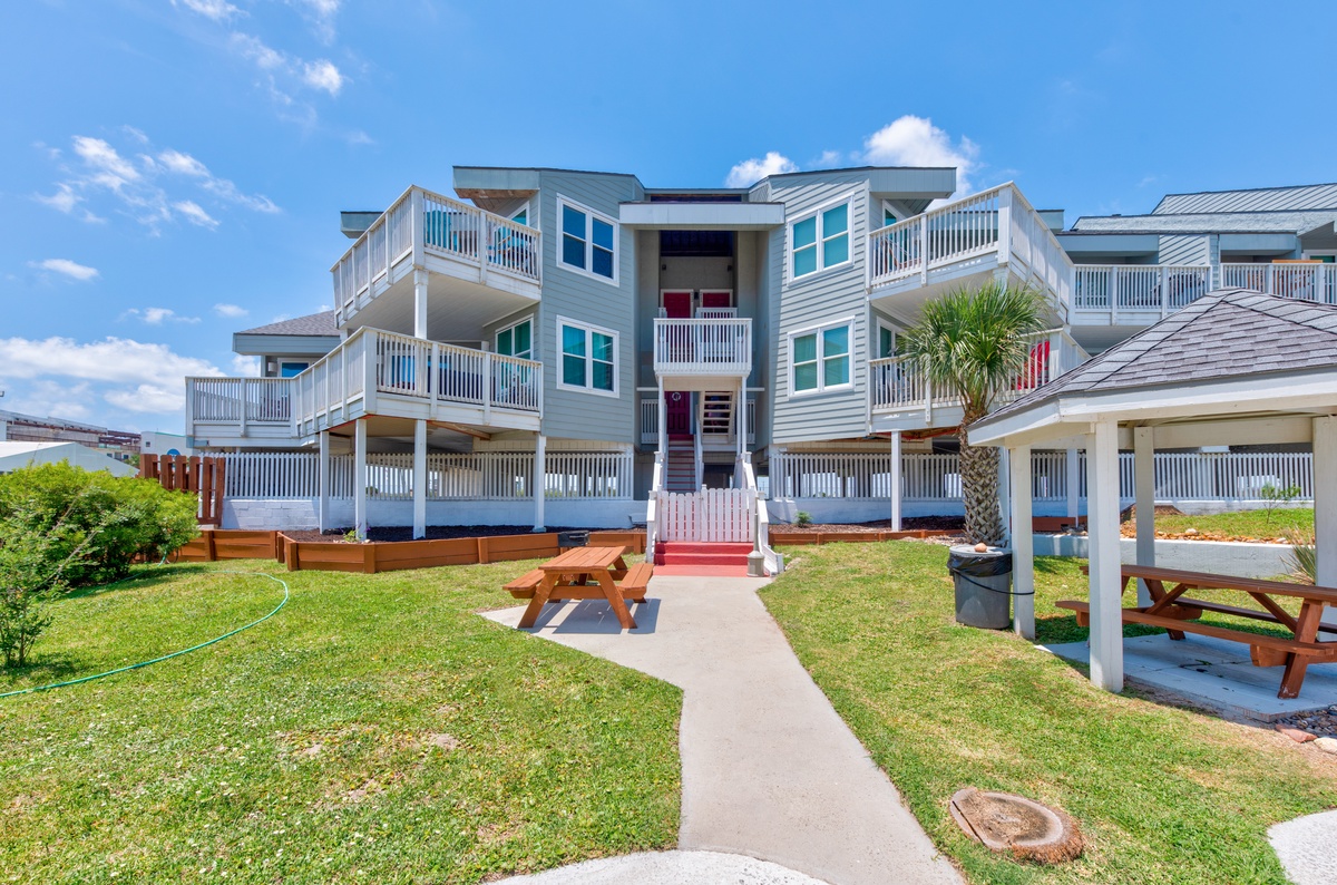 Modern beachside property featuring multiple balconies and outdoor gathering spaces with picnic tables in a sunny coastal setting.