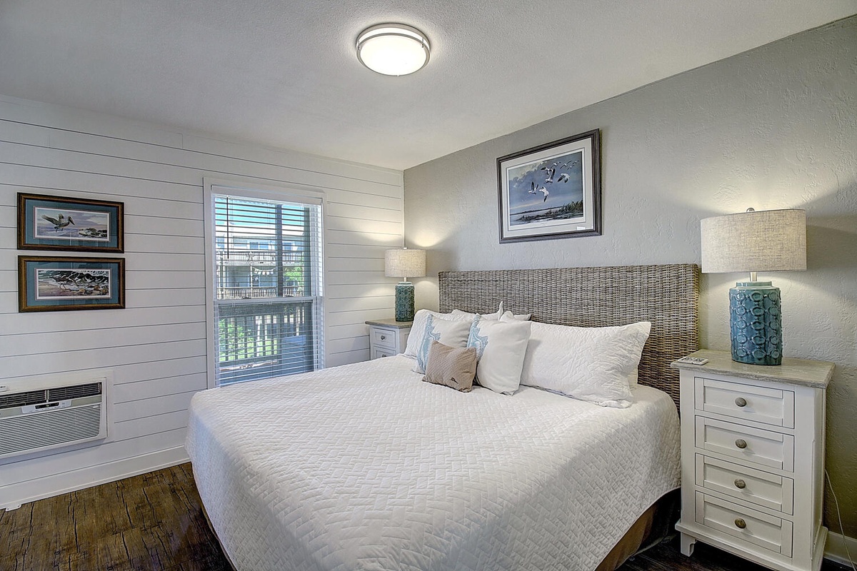Sink into your plush bed beneath coastal artwork and soft lighting, where wicker textures and serene blues create a peaceful retreat.
