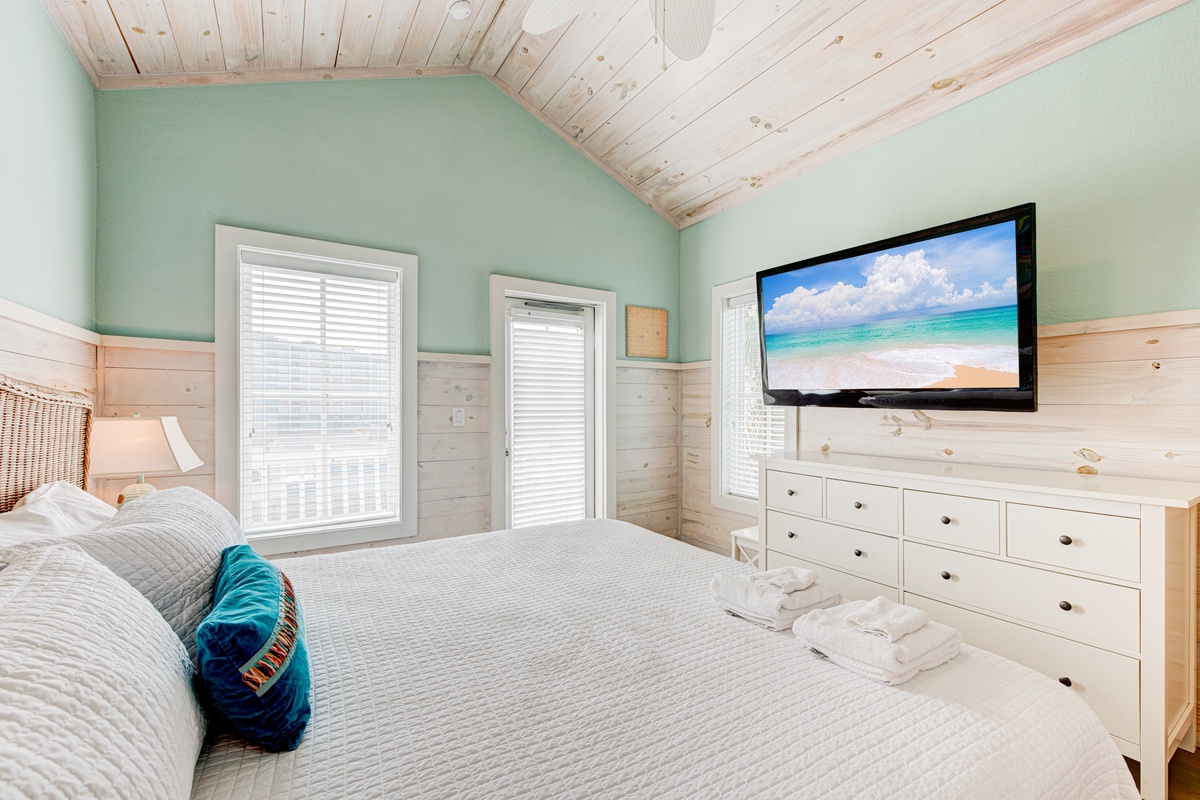 Relax in your coastal-inspired bedroom with soft mint walls, natural wood accents, and a large TV for unwinding after beach days.