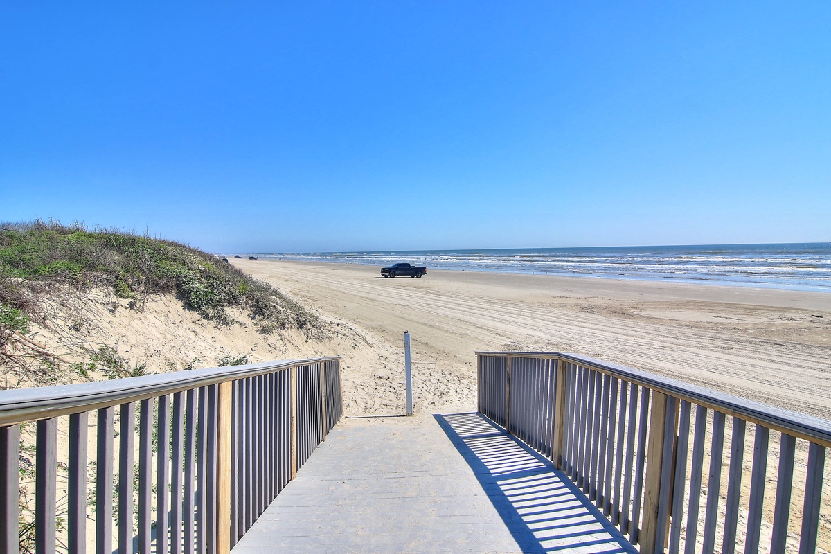 Wooden boardwalk leads directly to expansive sandy beach with clear blue skies and rolling waves.