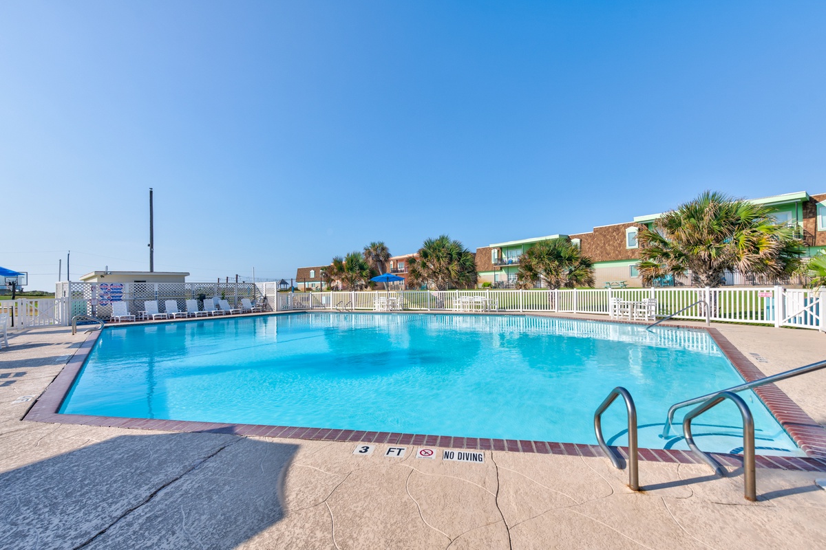 Large swimming pool surrounded by tropical palms and colorful beachside buildings under clear blue skies.