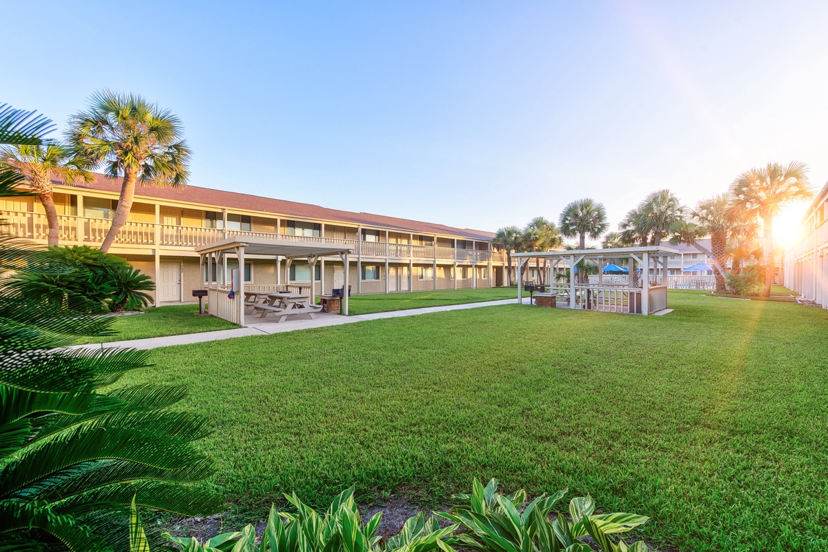 Tropical resort complex featuring lush palm trees, manicured lawns, and multiple buildings with covered balconies under golden sunset lighting.