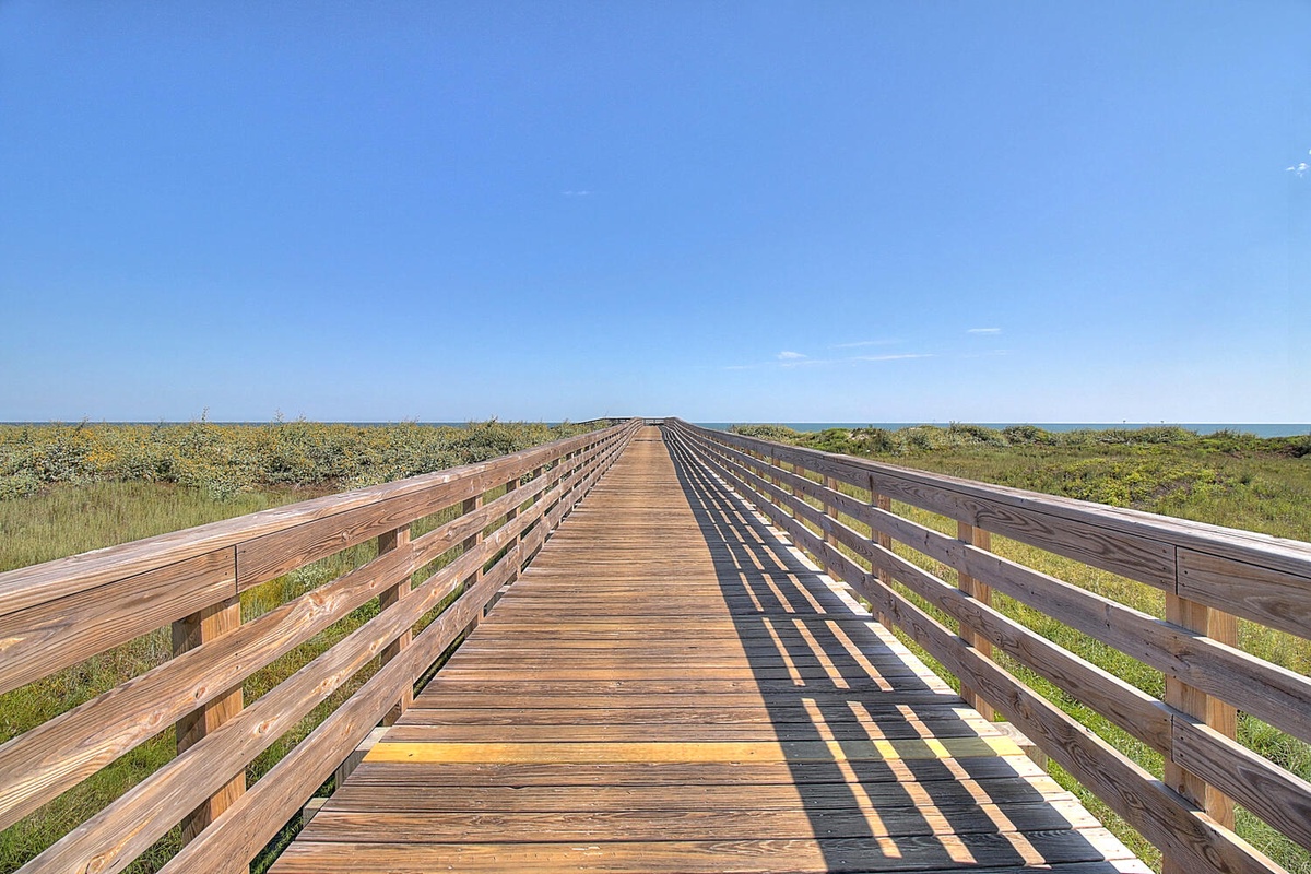 A wooden boardwalk stretches through natural coastal landscape under bright blue skies, perfect for peaceful walks and nature exploration.
