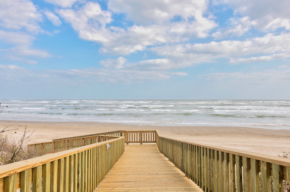 Wooden boardwalk leads to pristine sandy beach with rolling waves under blue skies.