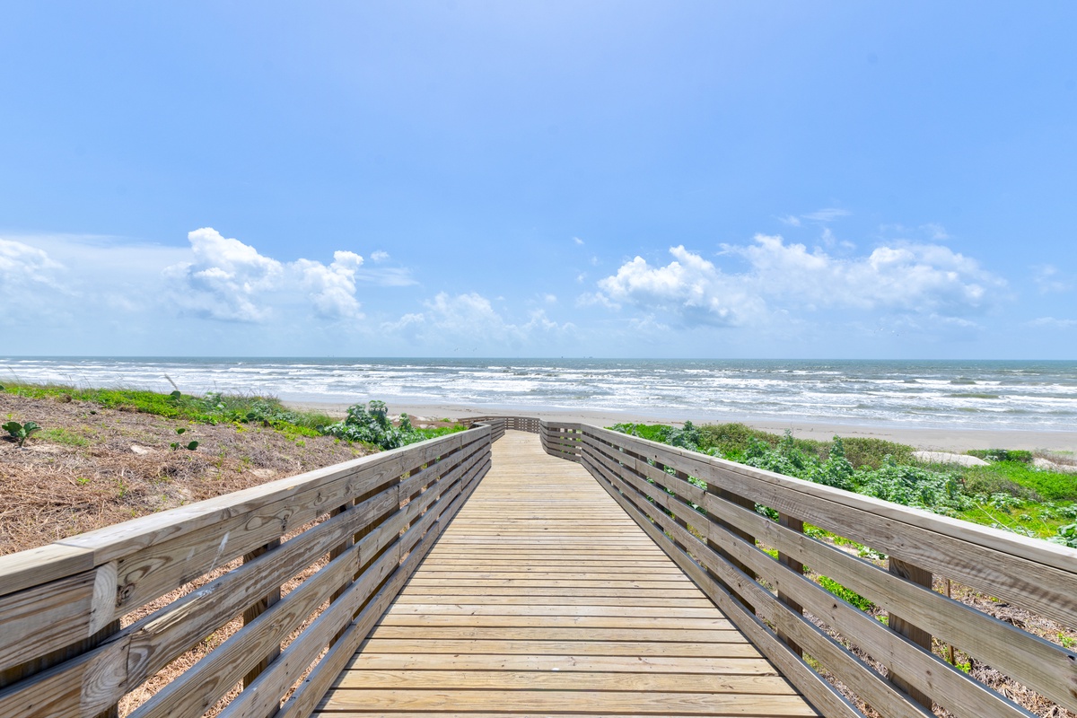 Wooden boardwalk leads across coastal dunes to pristine sandy beach with rolling waves under blue skies.