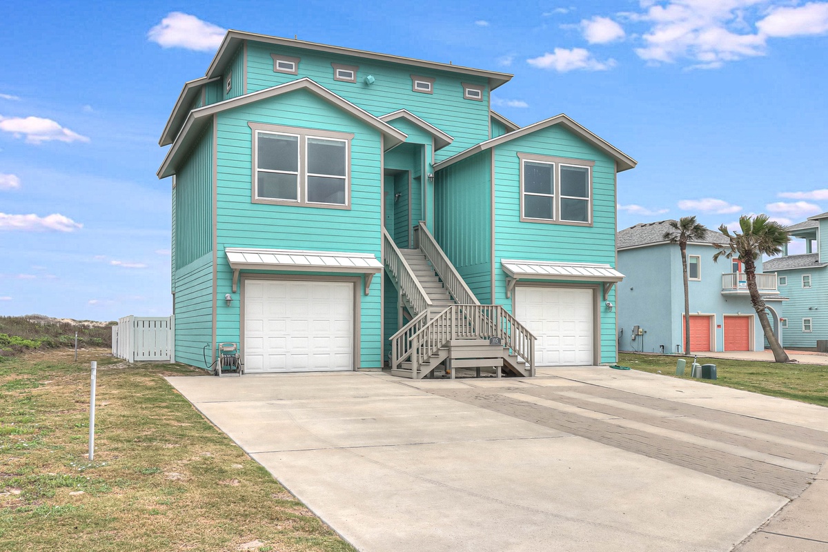Charming turquoise beach house with covered parking and staircase entrance, nestled among coastal palms in a peaceful neighborhood setting.