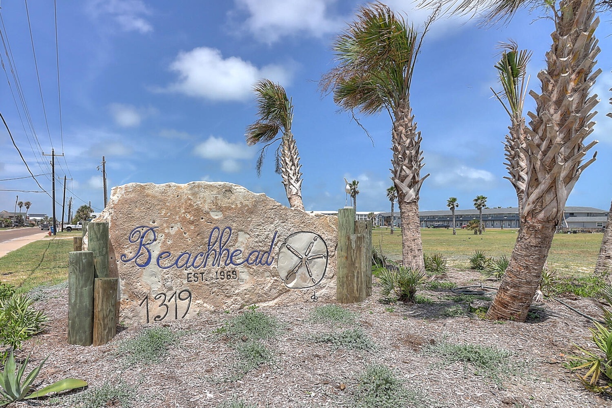 Coastal property entrance featuring decorative stone signage surrounded by palm trees in a beachside community setting.