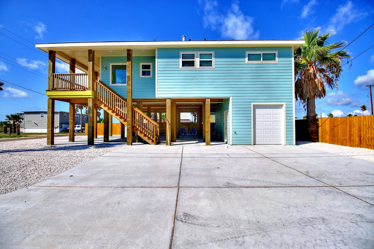Bright turquoise beach house on stilts with covered parking and palm trees under sunny blue skies.