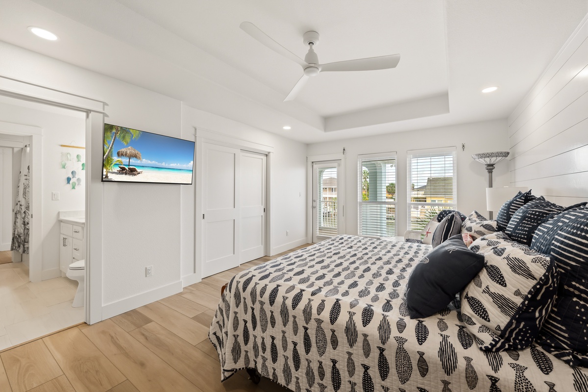 Your bright coastal bedroom features polished wood floors, ceiling fan, elegant patterned linens, and glass doors opening to a private balcony with neighborhood views.