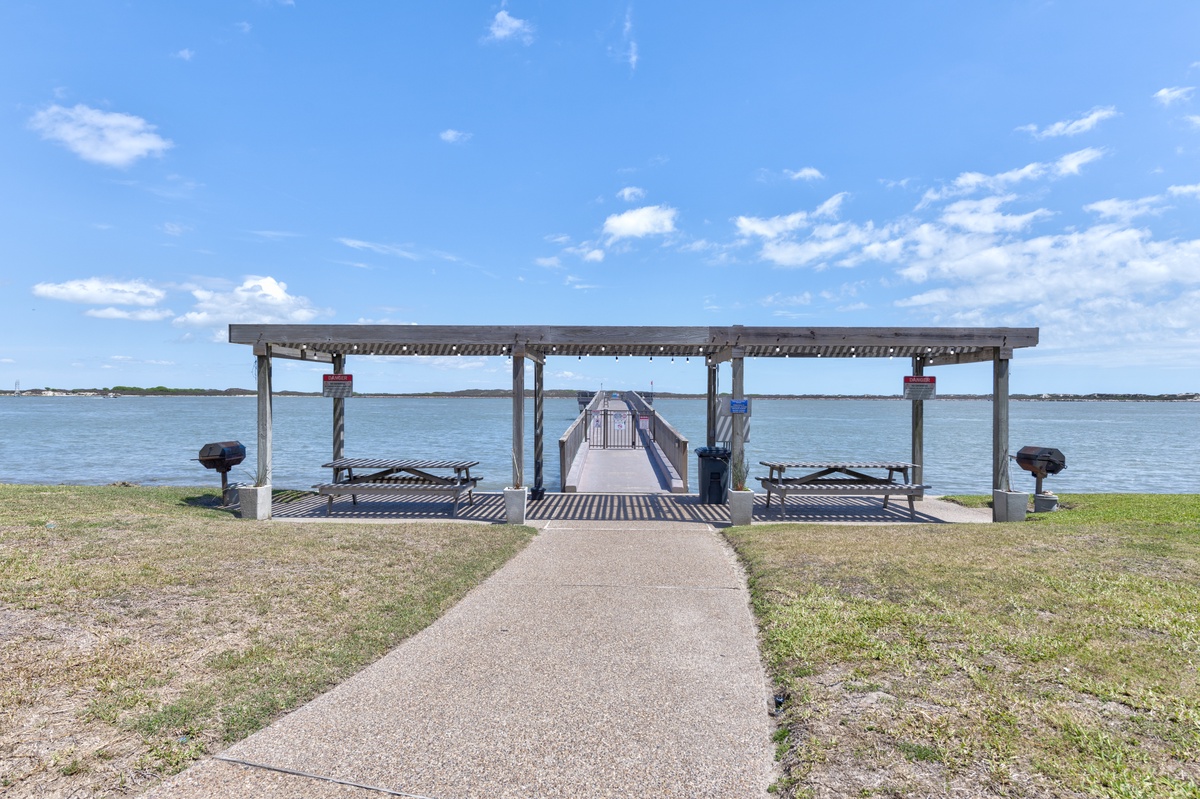 Waterfront pavilion with picnic tables and fishing pier overlooks calm bay waters under bright blue skies.