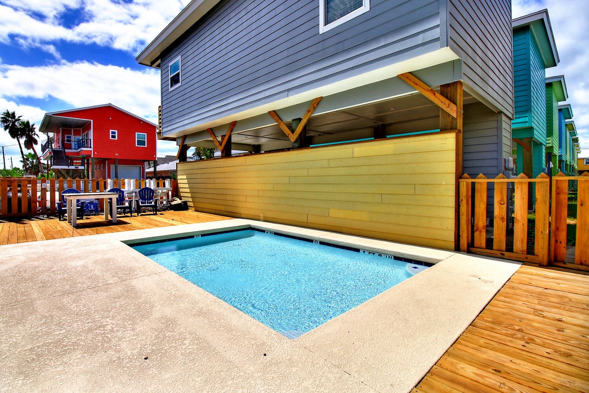 Dive into your private pool surrounded by colorful coastal homes, where wooden decking meets crystal-clear waters under bright blue skies.
