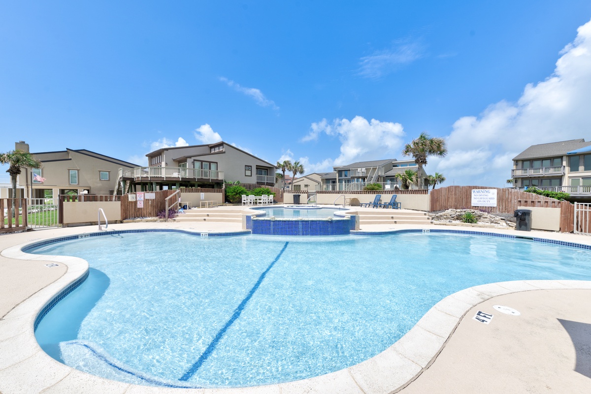 Community pool area surrounded by beach houses under clear blue skies with nearby hot tub and seating areas.