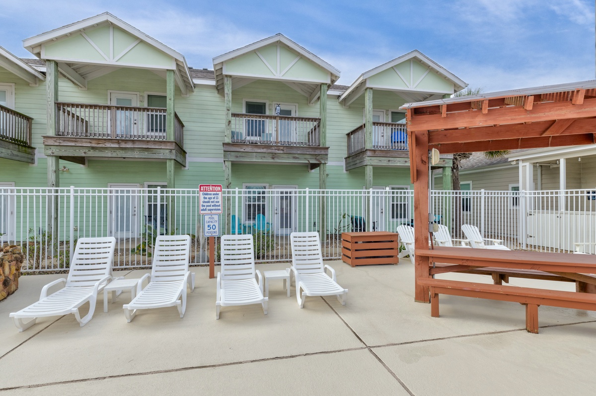Charming green coastal townhomes with private balconies, poolside lounging area with white chairs and wooden pergola for relaxation.