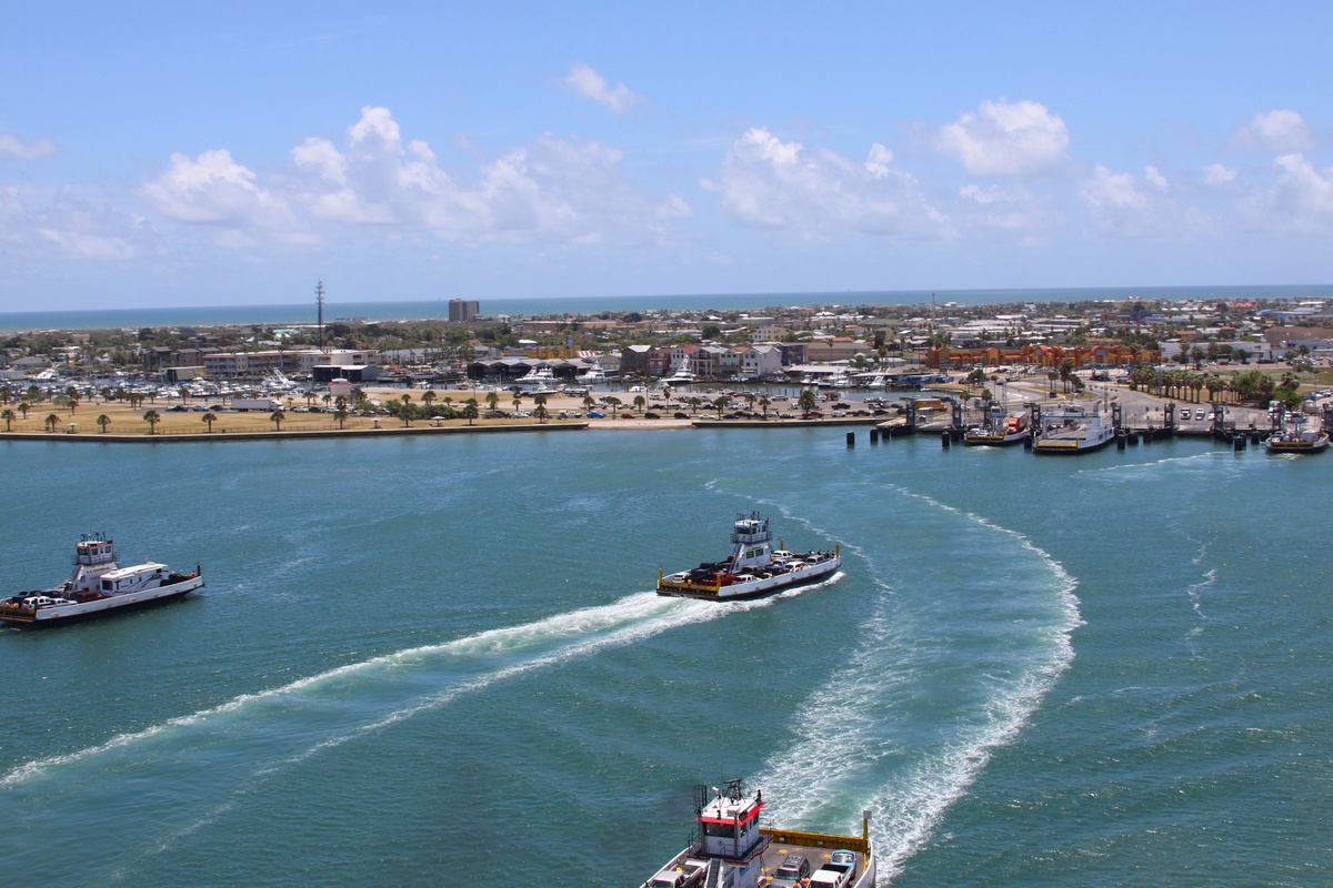 Aerial view of the bustling harbor with boats creating white wakes in turquoise waters and the coastal town stretching inland.