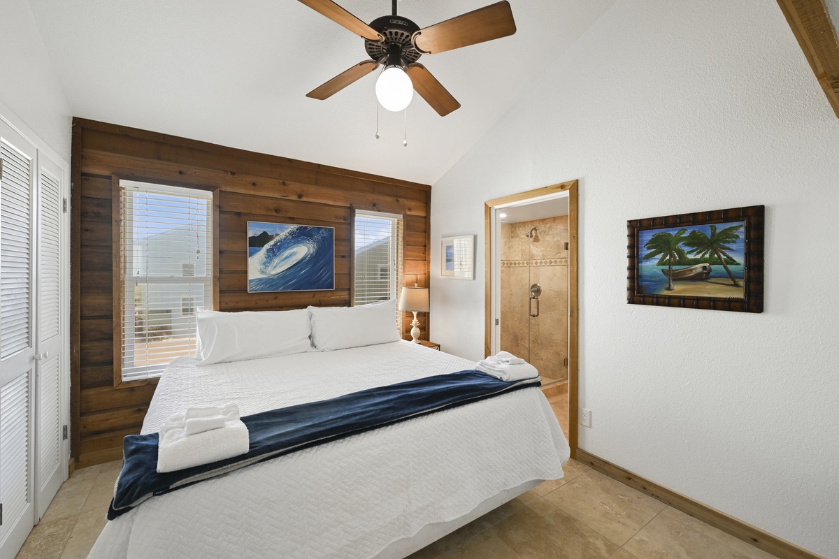 Drift off under vaulted ceilings and a ceiling fan in this cozy bedroom featuring a plush bed, warm wood accent wall, coastal artwork, and your own en-suite bath.