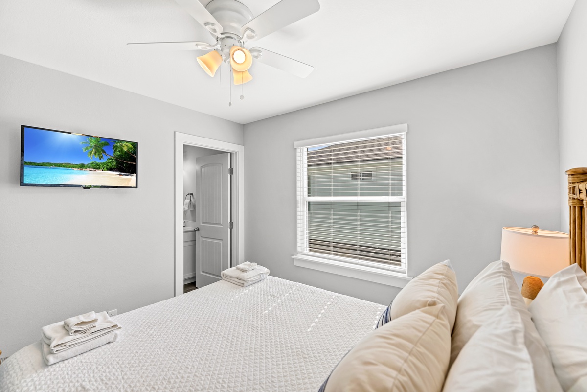 Sink into crisp white linens in your peaceful bedroom, where natural light filters through venetian blinds and tropical scenes on the wall-mounted TV await your relaxation.