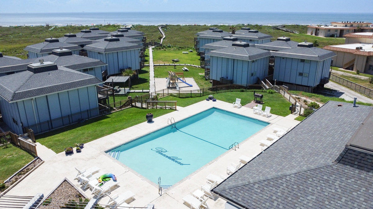 Aerial view of coastal vacation rental complex with swimming pool, playground, and beach access nearby.