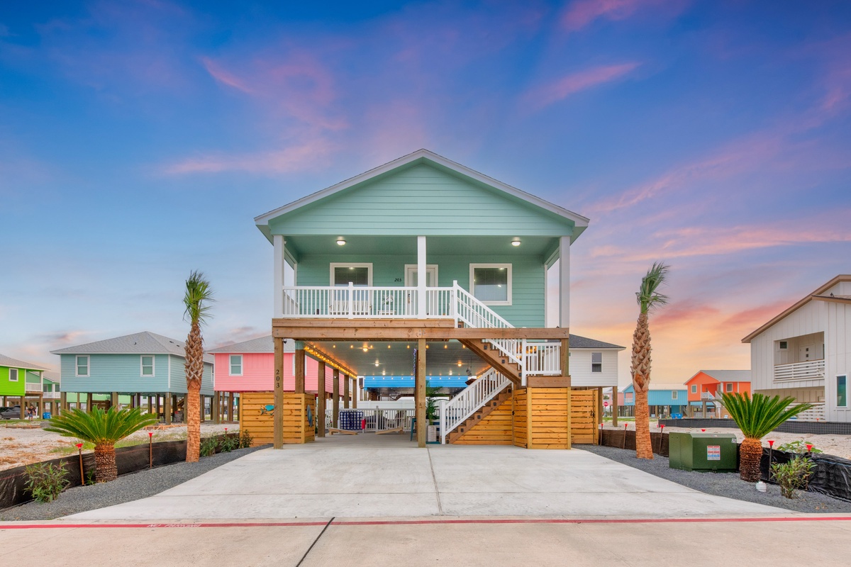 Charming beach house with elevated design and covered parking area, surrounded by colorful coastal homes under a painted sunset sky.