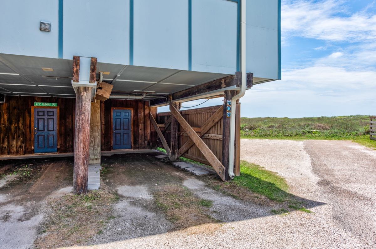 Covered parking shelter with rustic wooden gate and blue-trimmed building facade, surrounded by natural coastal landscape.