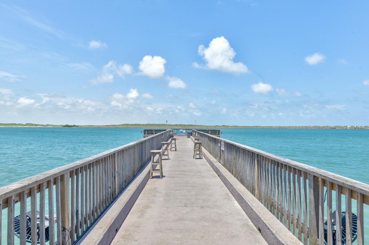 A scenic wooden pier extends into crystal-clear turquoise waters under brilliant blue skies with puffy white clouds.