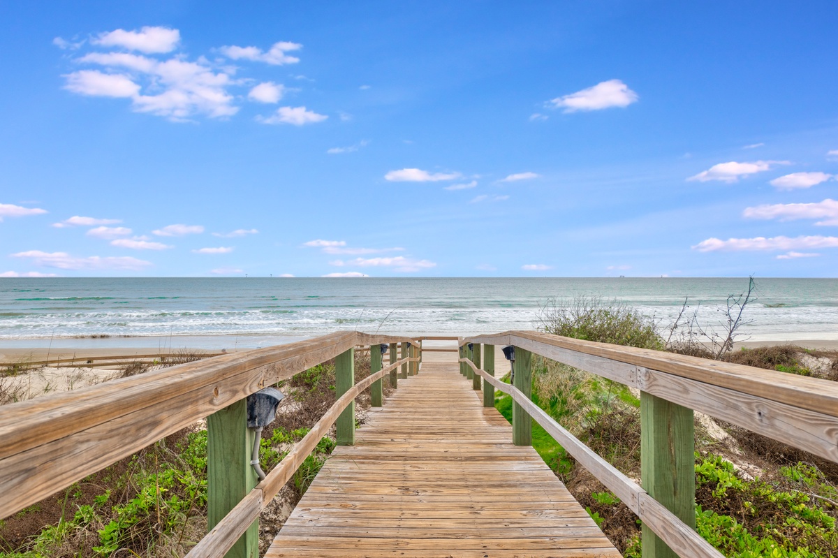 Wooden boardwalk leads directly to pristine beach with rolling waves and endless ocean views under blue sky.