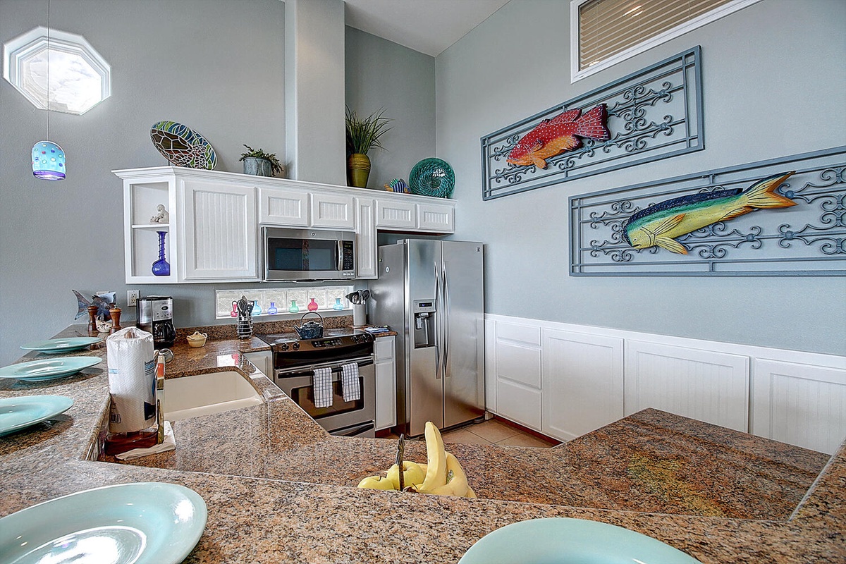 Your fully equipped kitchen features granite counters, stainless appliances, and coastal charm with vibrant fish artwork adorning the walls.