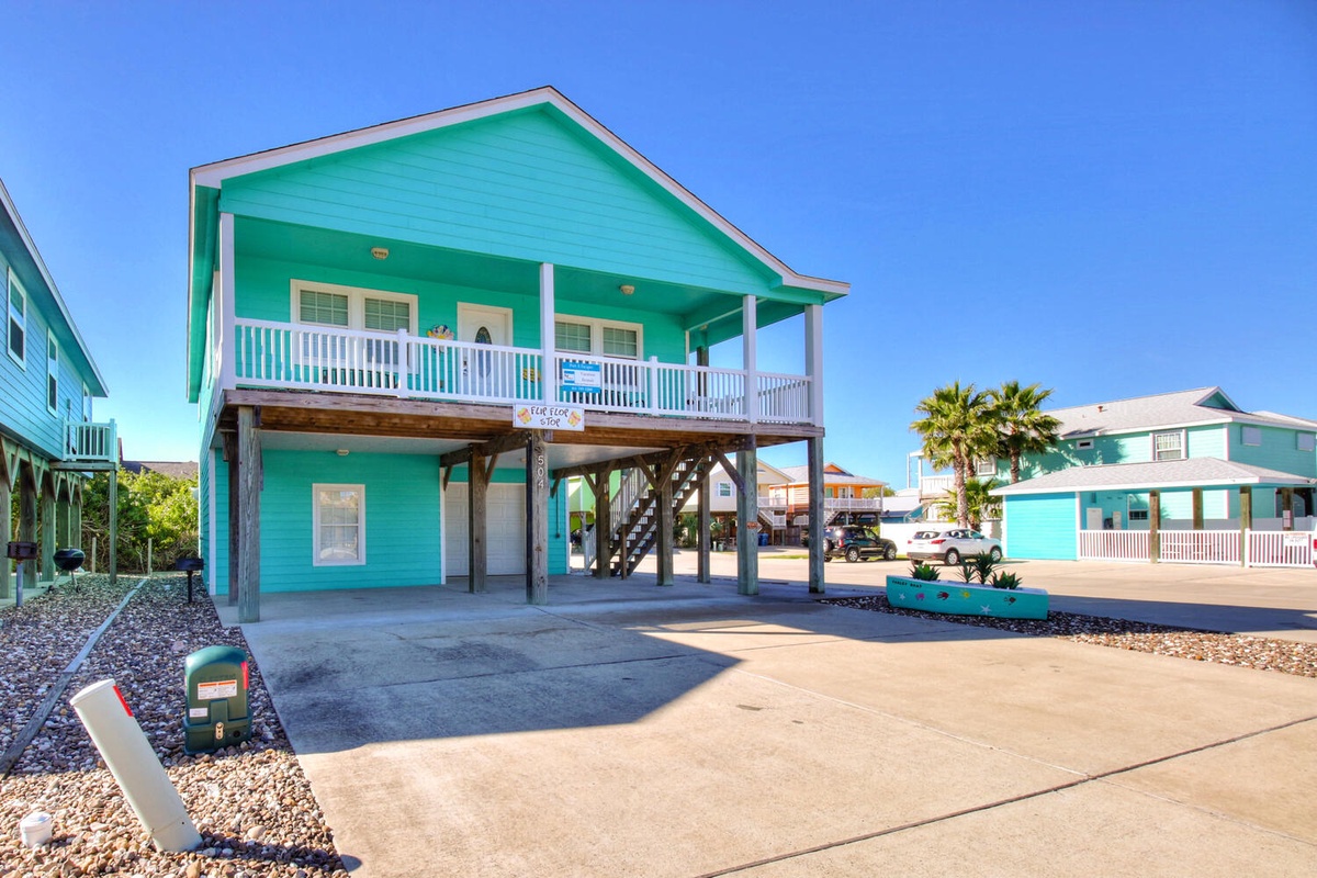 Vibrant turquoise beach house elevated on stilts with covered parking, white balcony railings, and tropical palm trees nearby.