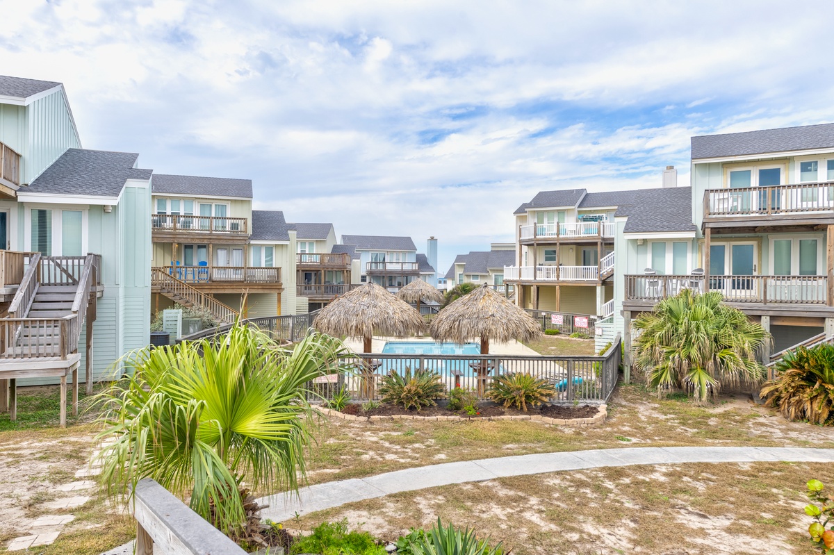 Coastal community featuring beach houses with balconies, tropical palms, thatched umbrellas, and a central swimming pool in a relaxed seaside setting.