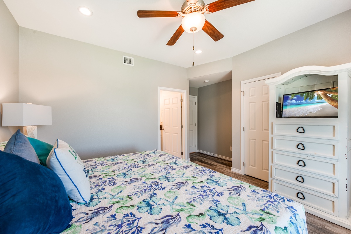 Unwind in your peaceful retreat with tropical-themed linens, entertainment center, and gentle ceiling fan for ultimate comfort.