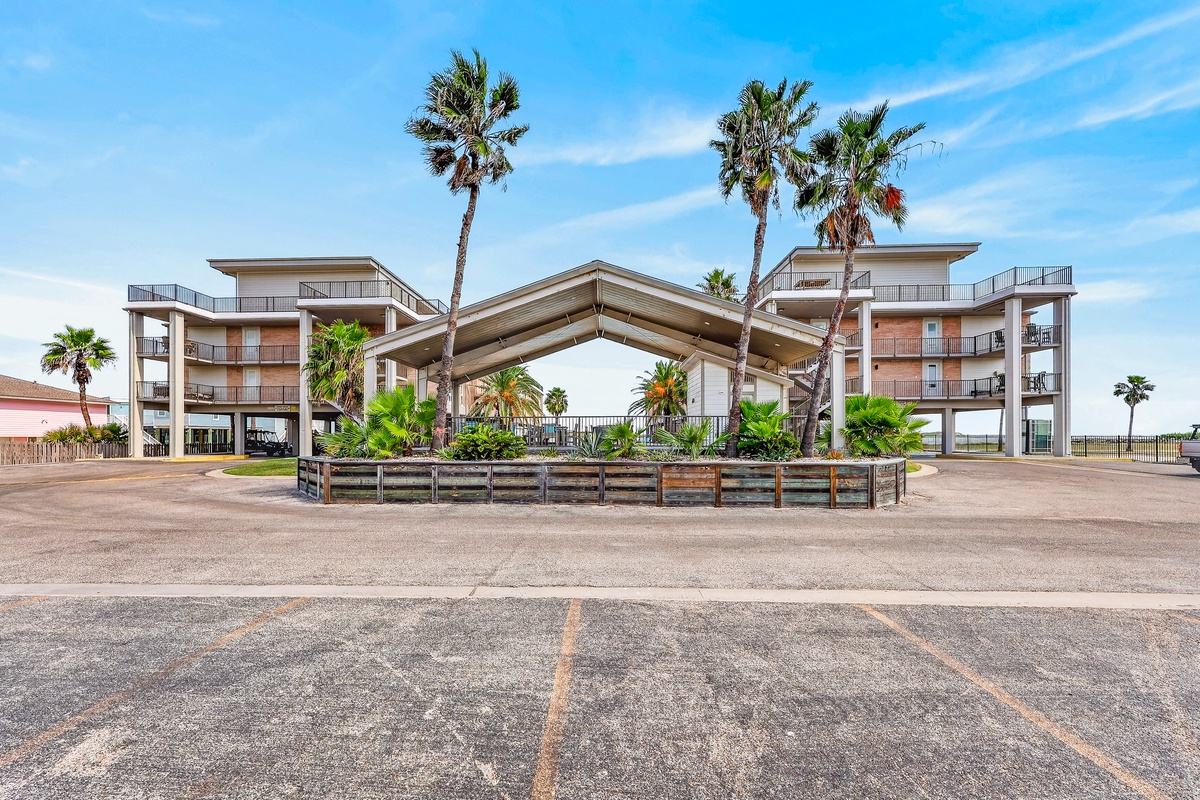 The property features a distinctive covered entrance framed by towering palm trees, with modern multi-story buildings set against a coastal backdrop.