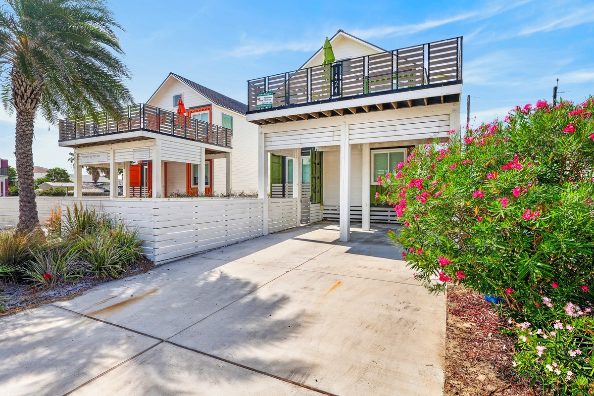 A modern beachside property featuring two-level accommodations with rooftop balconies, tropical landscaping with vibrant flowering shrubs, and palm trees that frame the coastal setting.
