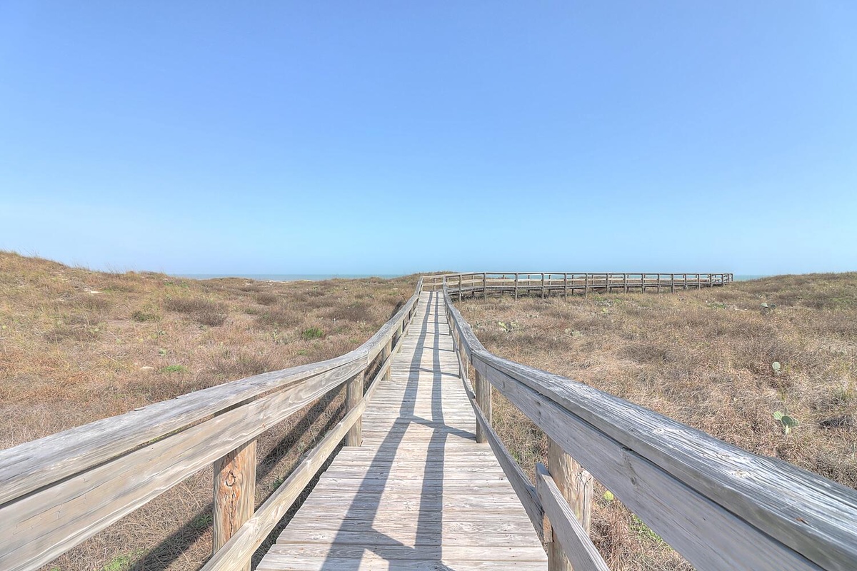 Wooden boardwalk winds through coastal landscape under clear blue skies, connecting to scenic viewpoint.