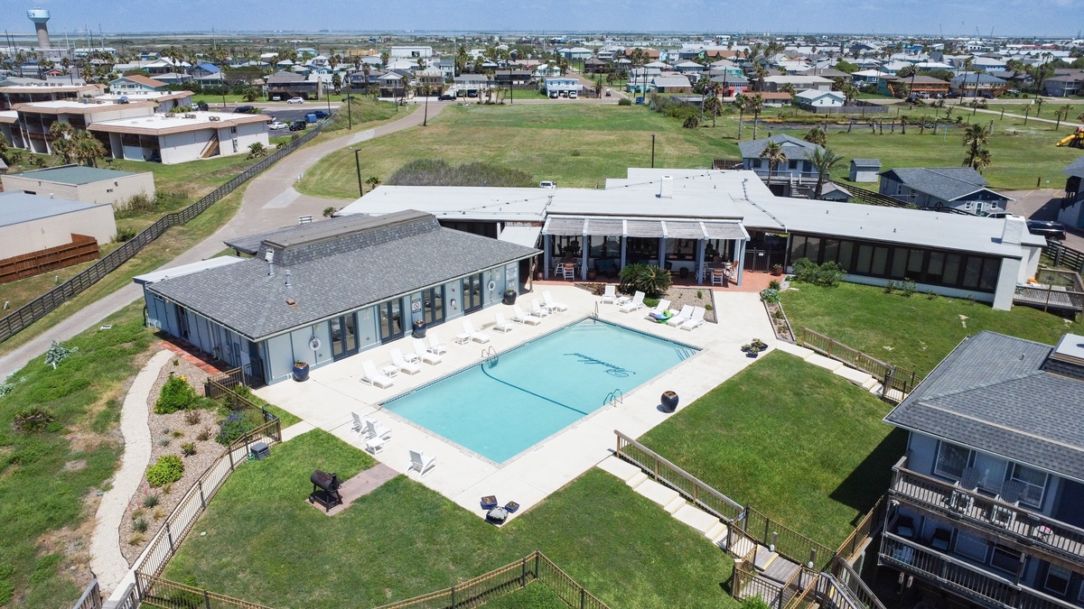 Modern coastal property featuring multiple buildings surrounding a pristine swimming pool with expansive green lawn areas and nearby residential neighborhood.