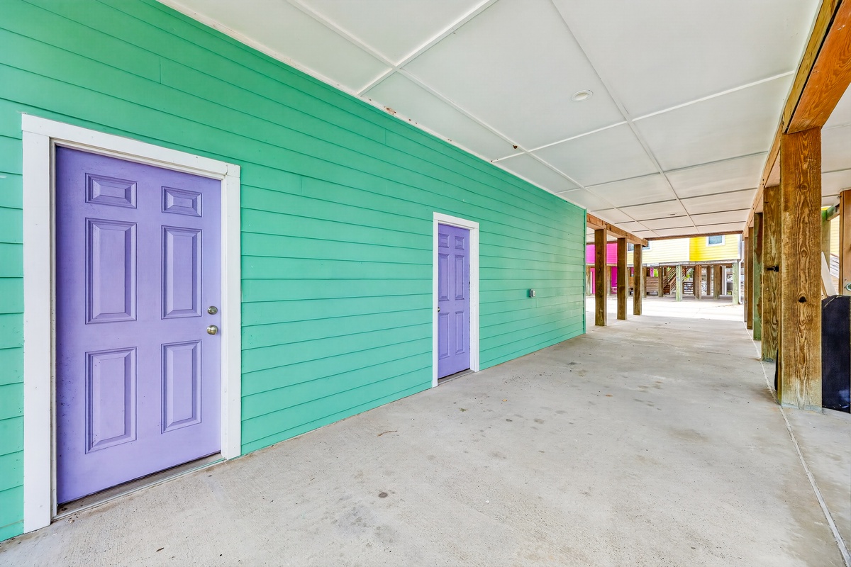 A covered walkway with vibrant turquoise walls and charming purple doors welcomes guests to their coastal retreat, offering sheltered access to individual units.