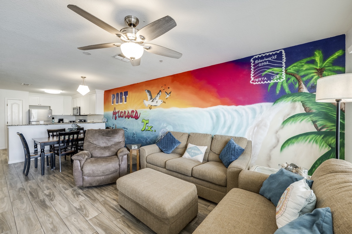 Unwind in your vibrant living space featuring a colorful Port Aransas beach mural, plush seating, and open-concept design connecting you to the fully equipped kitchen and dining area.