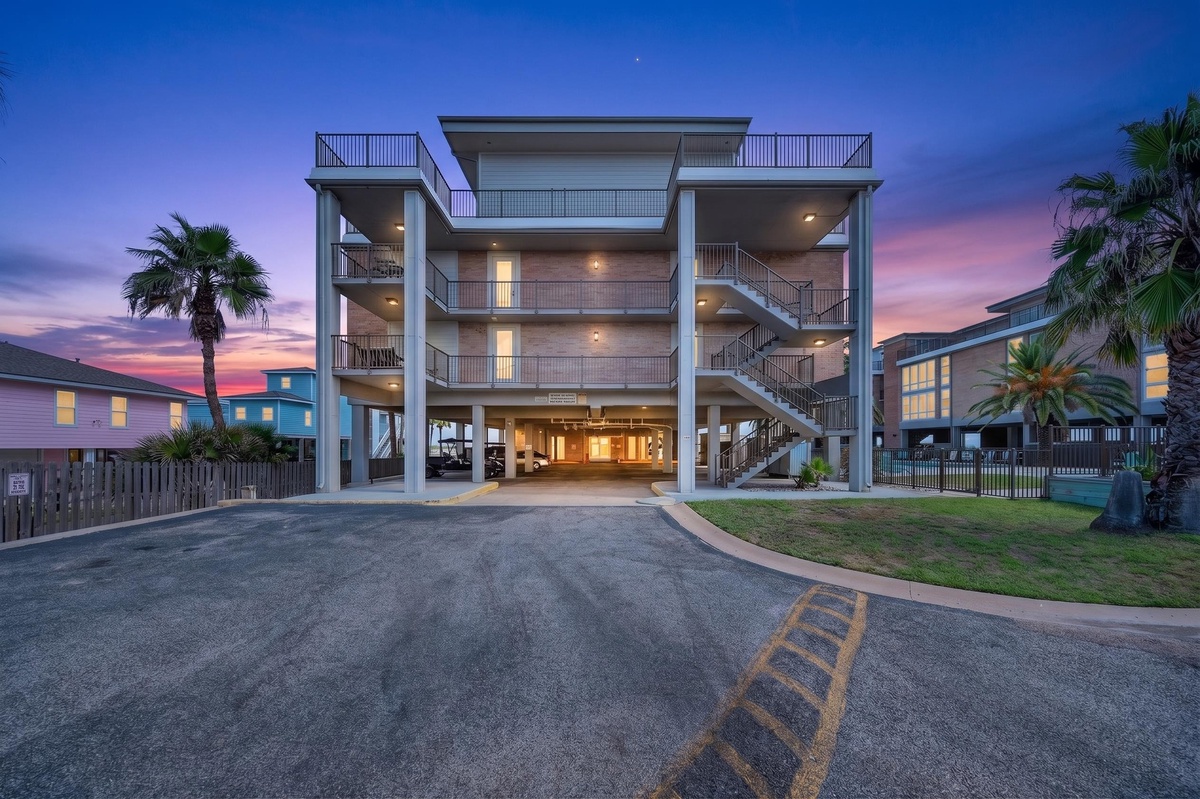 Modern beachside property with palm-fringed grounds and spacious covered parking beneath multiple levels, framed by a stunning coastal sunset.